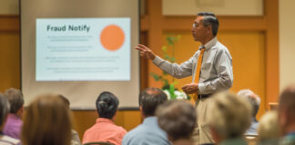 REALTOR® leading a community workshop on deed fraud prevention in a local hall, pointing to a presentation on 'Fraud Notify'.