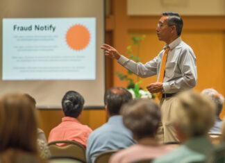 REALTOR® leading a community workshop on deed fraud prevention in a local hall, pointing to a presentation on 'Fraud Notify'.